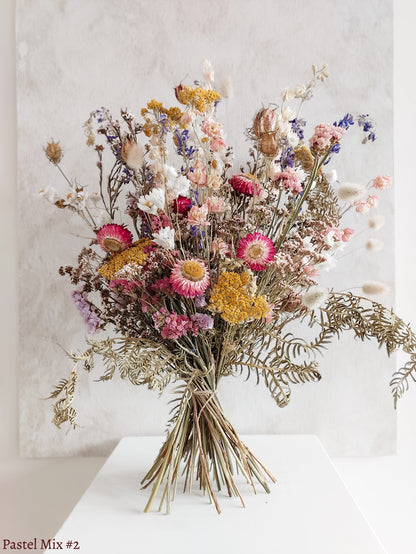 A dried flower bouquet featuring soft pink, yellow, and mauve flowers with soft green foliage, displayed on a white plinth against a textured gray background.