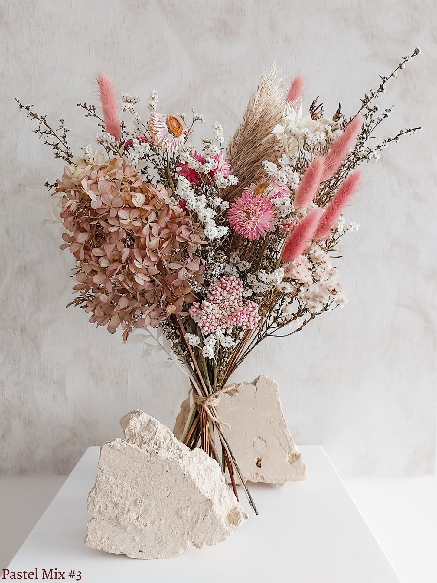 A dried flower bouquet featuring soft pink, white, and cream flowers against a textured light gray background.