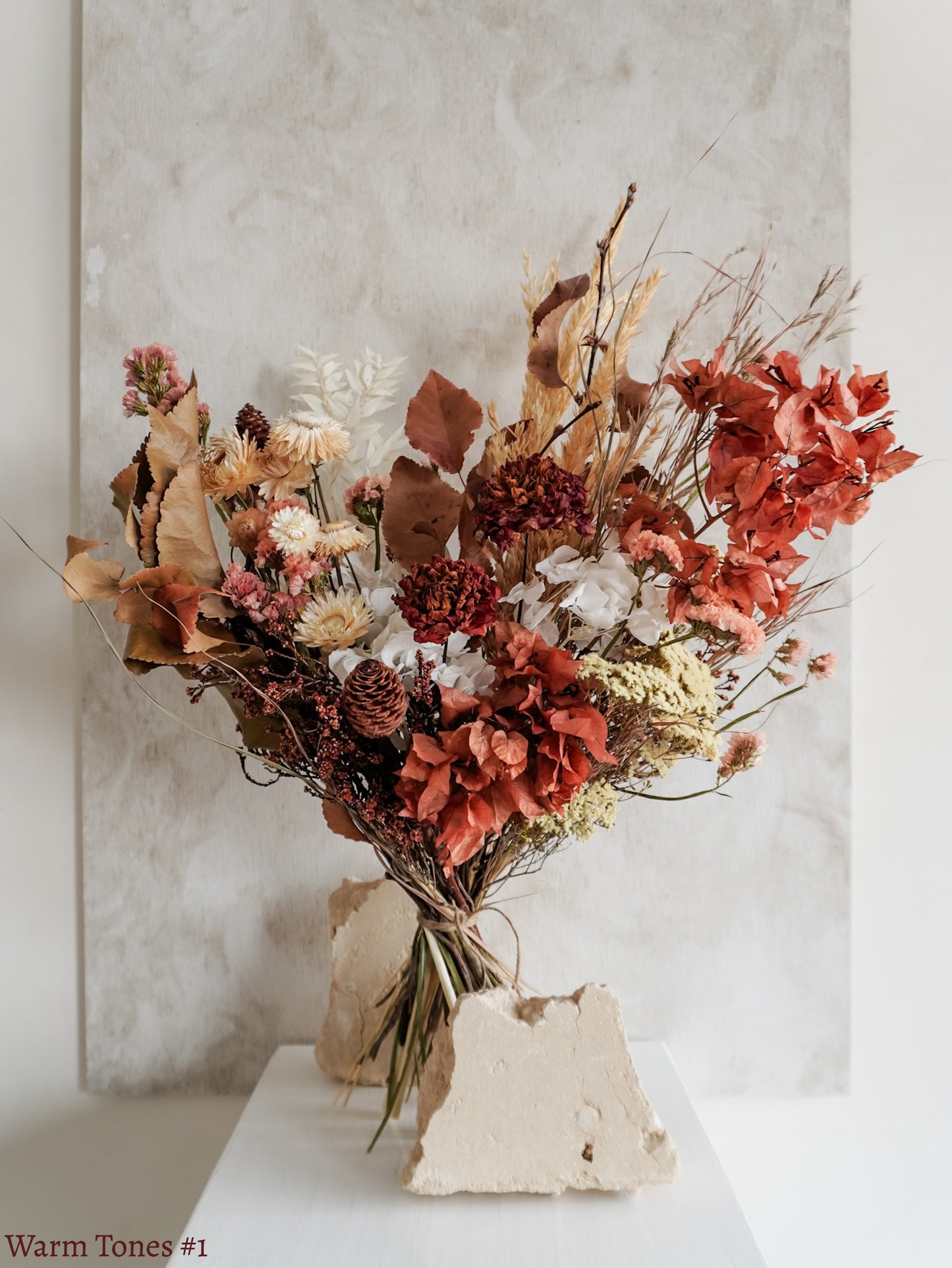 A dried flower bouquet featuring red, rust, and autumnal style flowers with earthy foliage, displayed on a white plinth against a textured gray background.