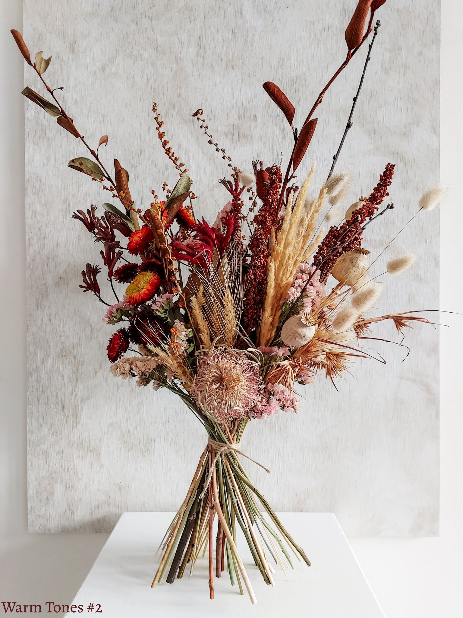 A dried flower bouquet featuring rich red, autumnal style flowers with earthy foliage, displayed on a white plinth against a textured gray background.