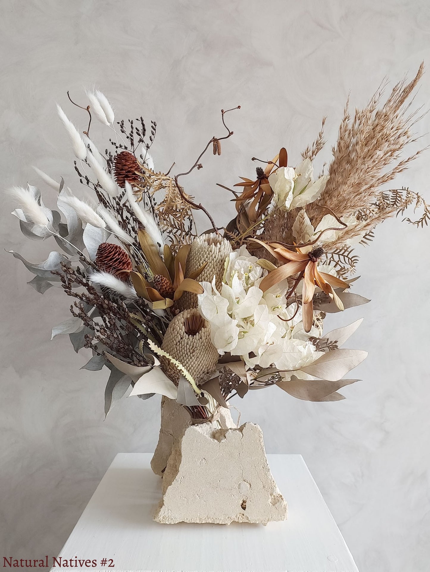 A dried flower bouquet featuring textural native flowers in natural earthy tones, displayed on a white plinth against a textured gray background.