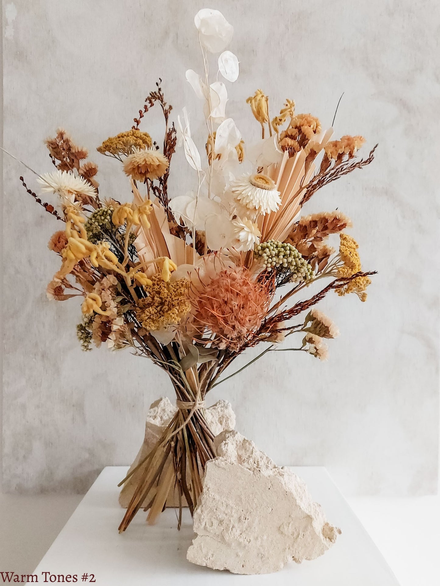 A dried flower bouquet featuring yellow, orange, and white flowers against a textured light gray background.