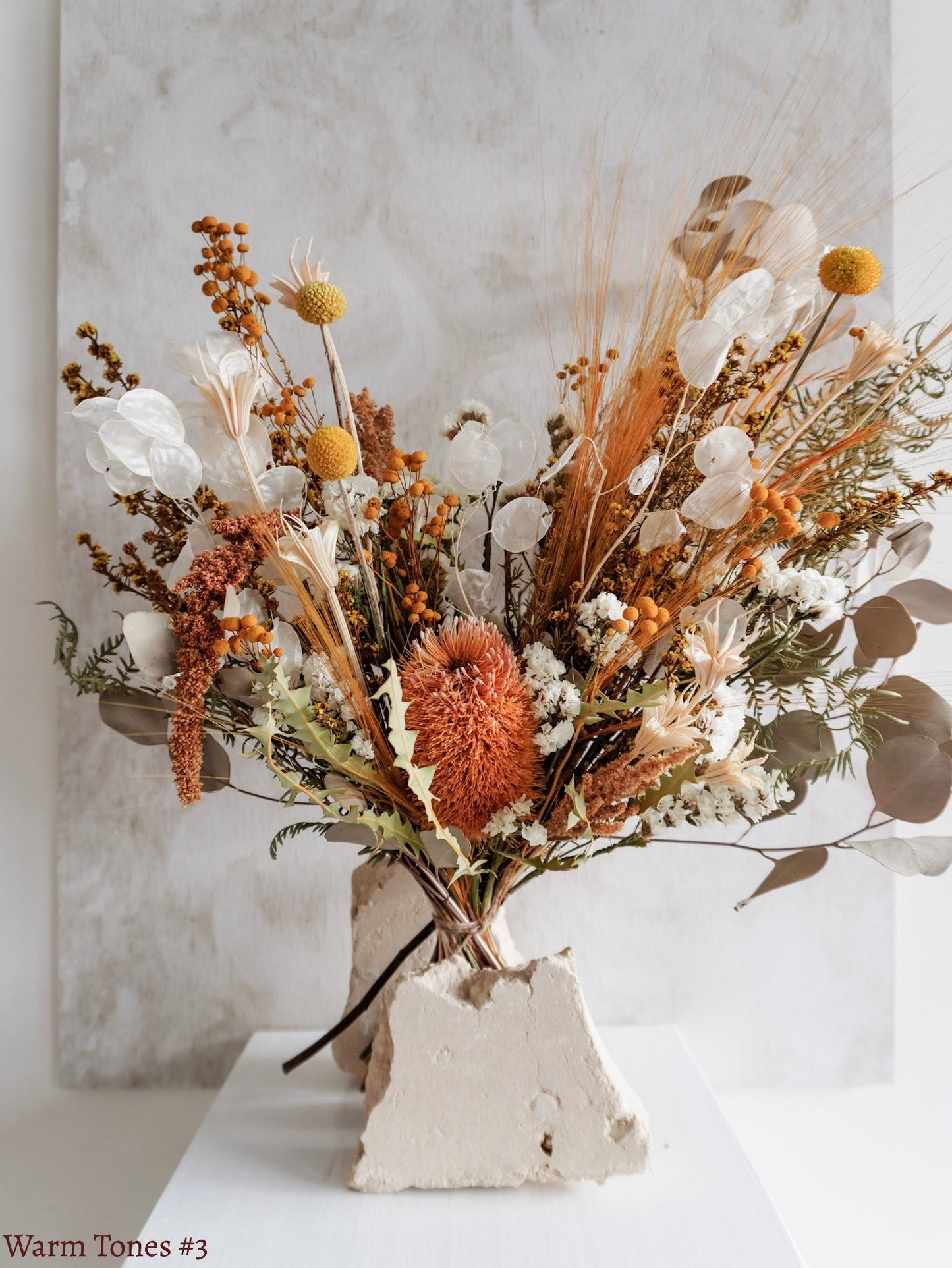 A dried flower bouquet featuring vibrant orange, yellow, and white flowers with soft green foliage, displayed on a white plinth against a textured gray background.