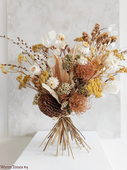 A dried flower bouquet featuring vibrant orange, yellow, and white flowers, displayed on a white plinth against a textured gray background.