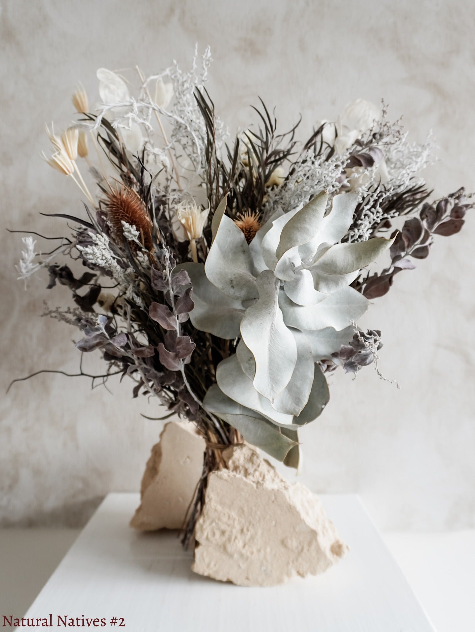 A dried flower bouquet featuring textural native flowers in silver and white tones against a textured light gray background.