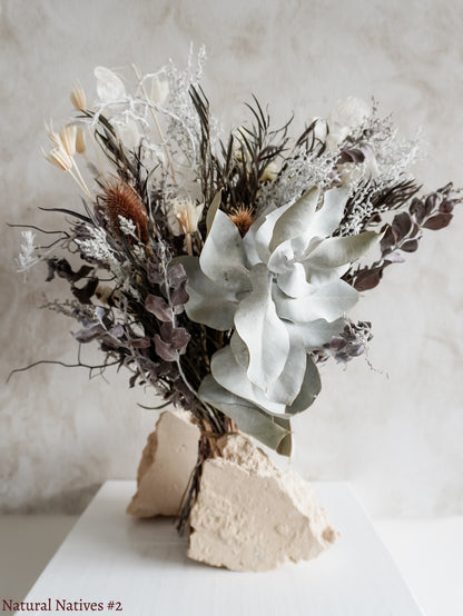 A dried flower bouquet featuring textural native flowers in silver and white tones against a textured light gray background.