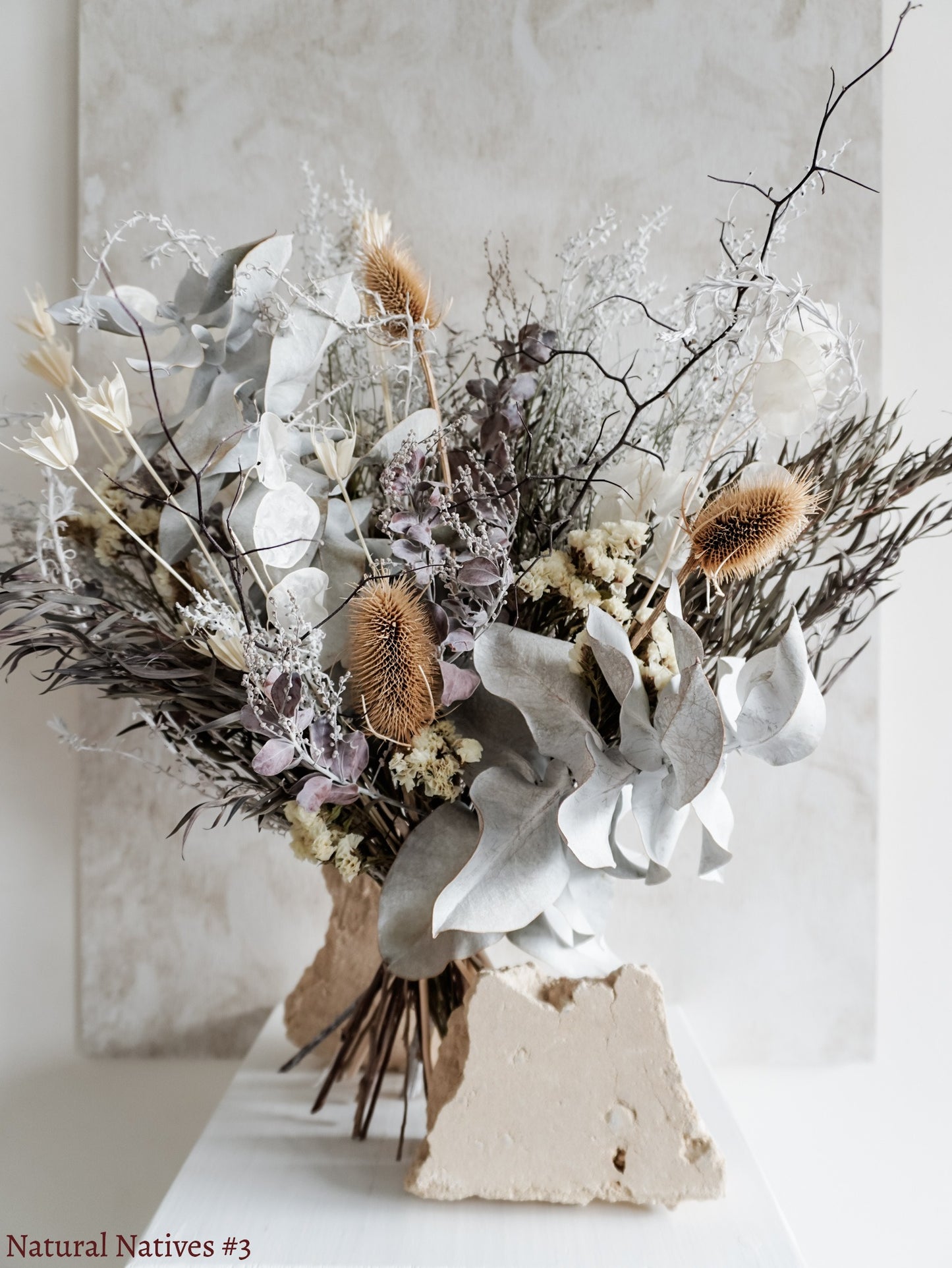 A dried flower bouquet featuring textural native flowers and foliage in wintery silver tones, displayed on a white plinth against a textured gray background.