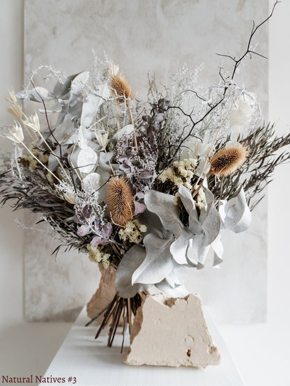 A dried flower bouquet featuring textural native flowers and foliage in wintery silver tones, displayed on a white plinth against a textured gray background.