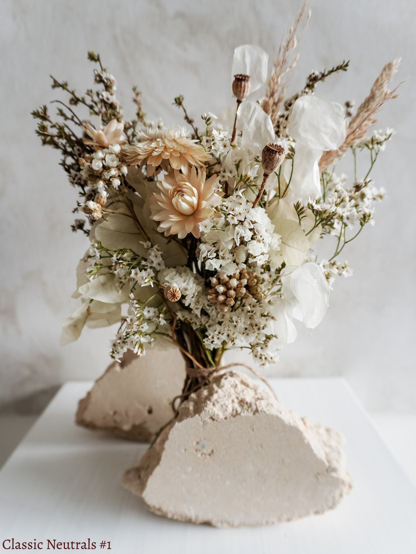 A petite dried flower bouquet featuring white and neutral tone flowers on a white plinth against a light gray background.