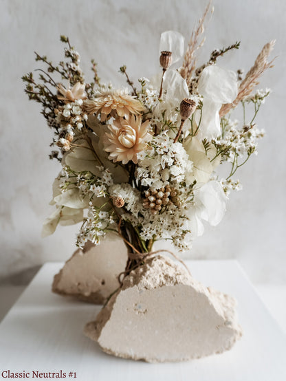 A petite dried flower bouquet featuring white and neutral tone flowers on a white plinth against a light gray background.