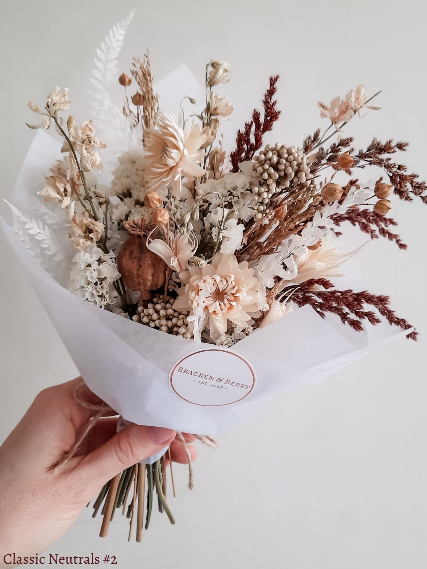 A petite dried flower bouquet wrapped in white tissue paper, held by a hand against a neutral background. The bouquet features white, cream, and earthy tone flowers.