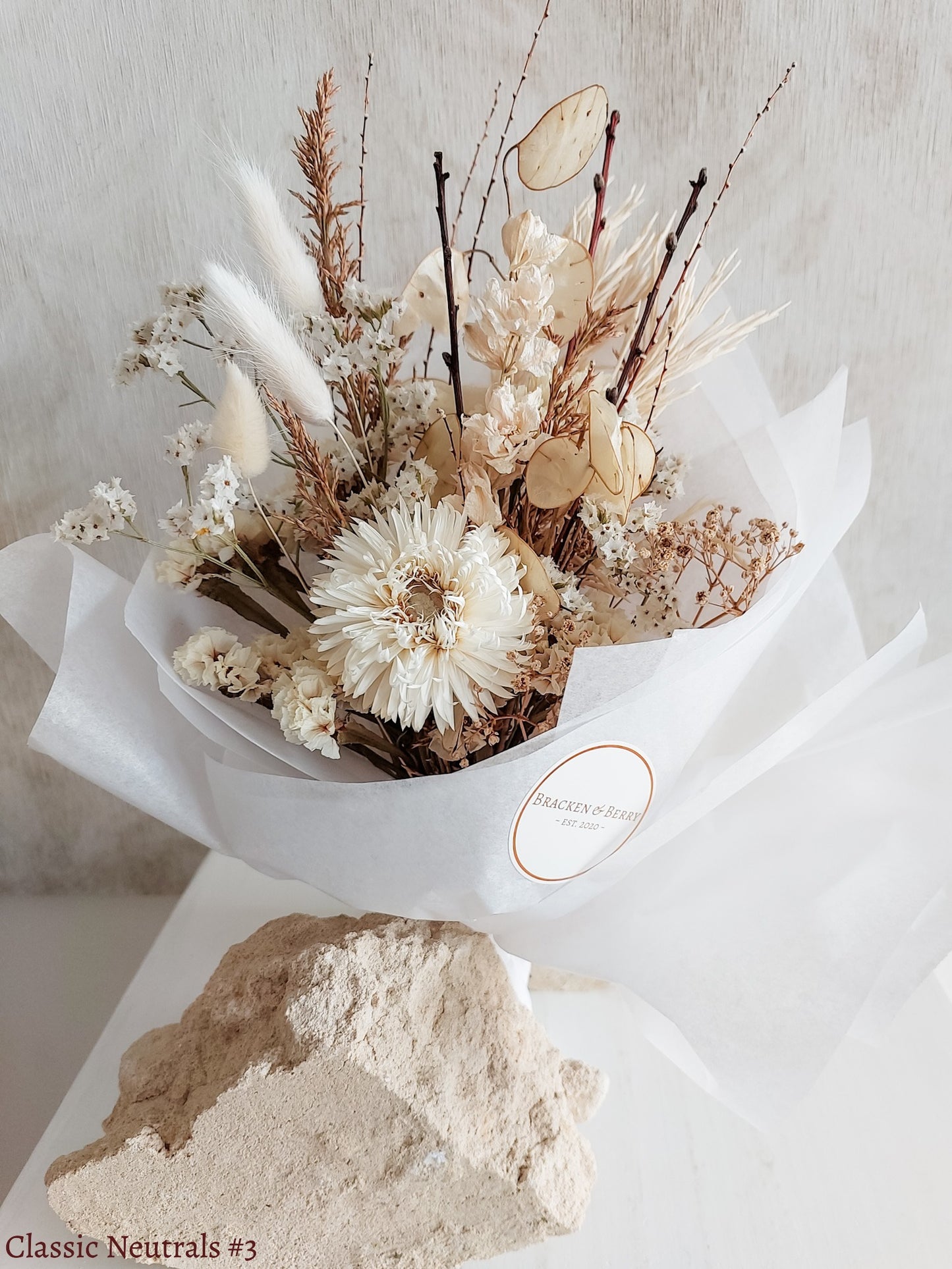 A petite dried flower bouquet wrapped in white tissue paper, featuring white and neutral tone flowers on a white plinth against a light gray background.