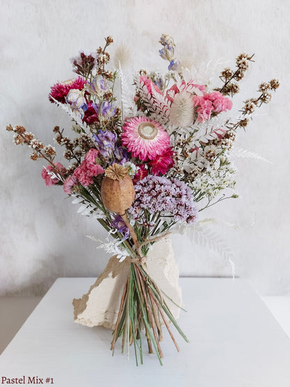 A petite dried flower bouquet featuring soft pink and mauve flowers on a white plinth against a light gray background.
