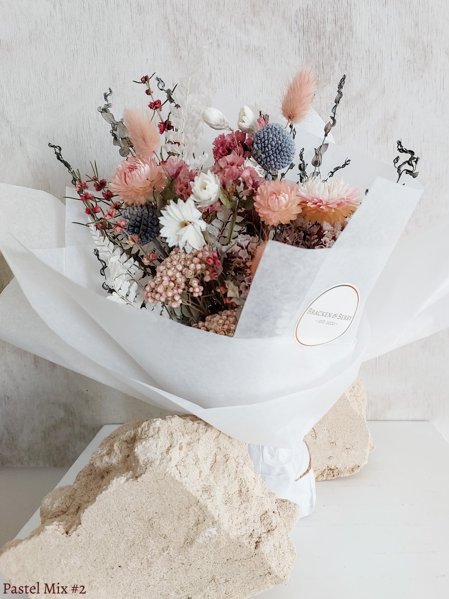 A petite dried flower bouquet wrapped in white tissue paper, featuring white, soft pink, and light blue flowers on a white plinth against a light gray background.