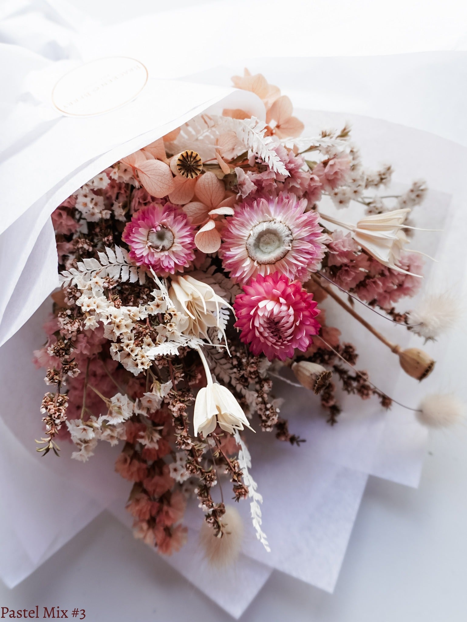 A petite dried flower bouquet wrapped in white tissue paper, featuring white, soft pink, and peach flowers laid flat on a white table.
