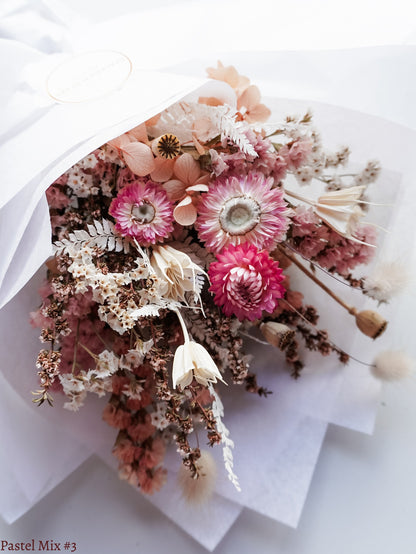 A petite dried flower bouquet wrapped in white tissue paper, featuring white, soft pink, and peach flowers laid flat on a white table.