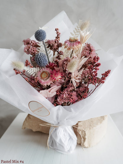 A petite dried flower bouquet wrapped in white tissue paper, featuring white, soft pink, and light blue flowers on a white plinth against a light gray background.
