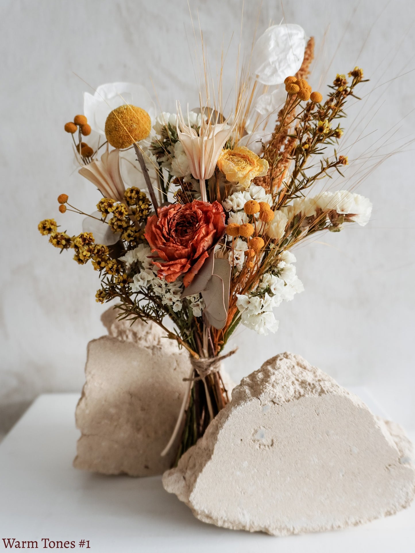 A petite dried flower bouquet featuring yellow, orange and white flowers on a white plinth against a light gray background.