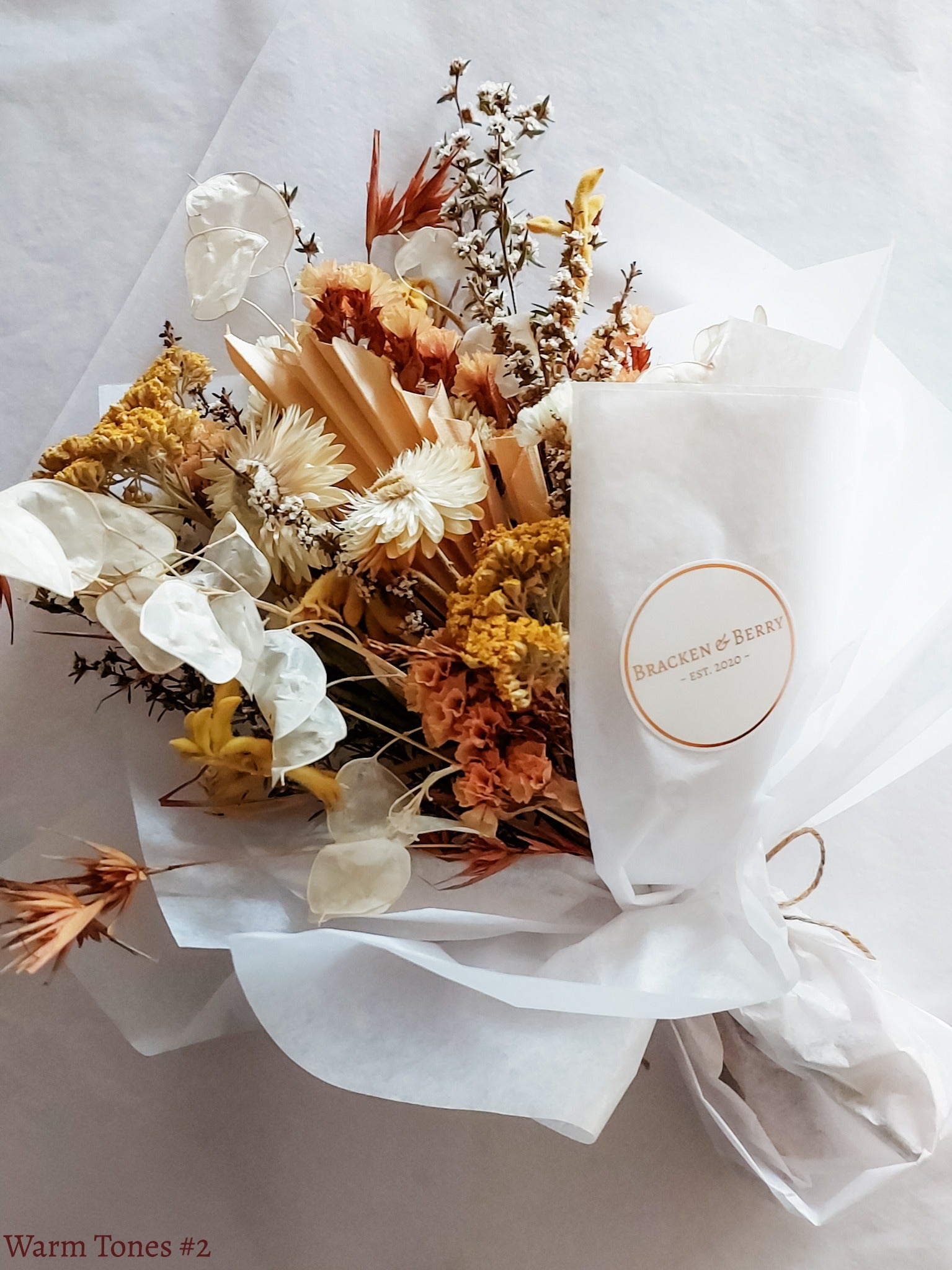 A petite dried flower bouquet wrapped in white tissue paper, featuring yellow, orange and white flowers laid flat on a white table.