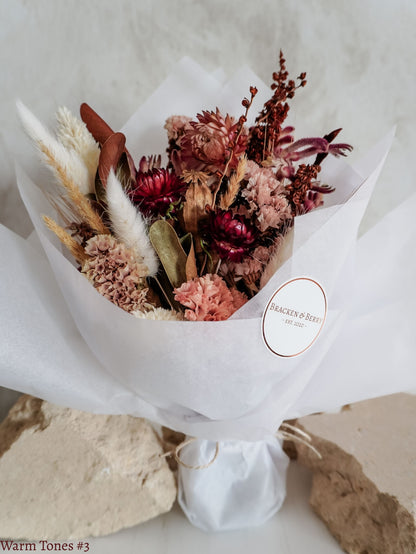A petite dried flower bouquet wrapped in white tissue paper, featuring autumnal tone flowers on a white plinth against a light gray background.