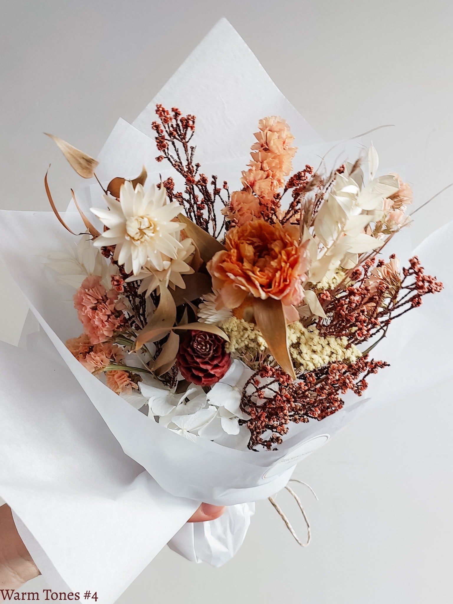 A petite dried flower bouquet wrapped in white tissue paper held by a hand against a neutral background. The bouquet features orange, white, and earthy tone flowers.