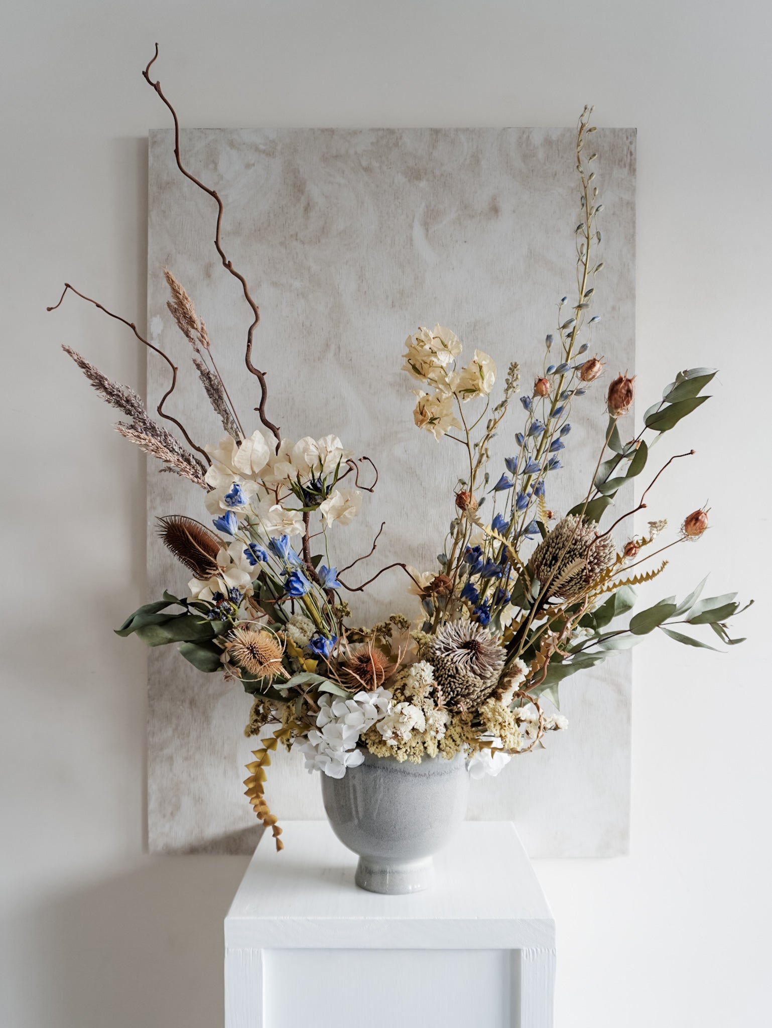Dried flower arrangement in ceramic vase with natural toned florals accented with blue delphinium – full view.