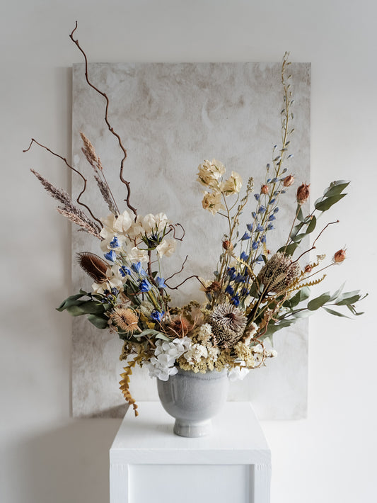 Dried flower arrangement in ceramic vase with natural toned florals accented with blue delphinium – full view.