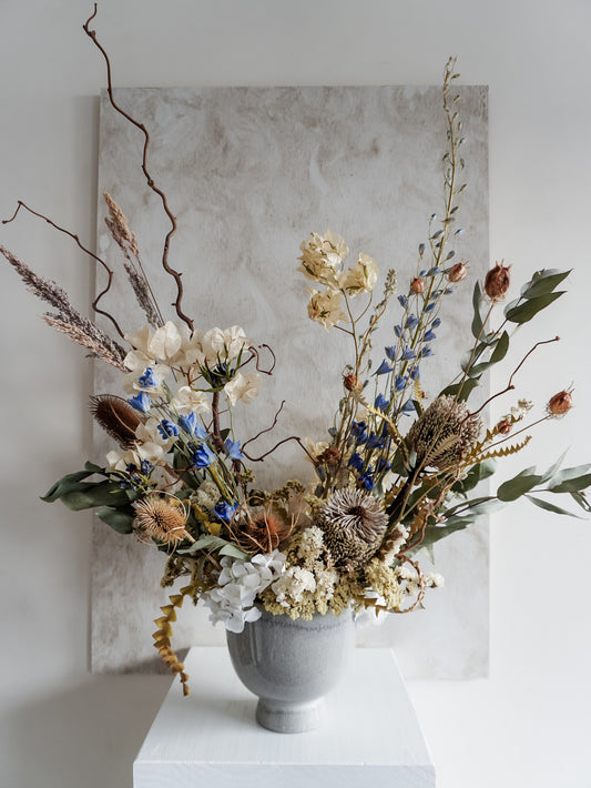 Dried flower arrangement in ceramic vase with natural toned florals accented with blue delphinium – close full view.