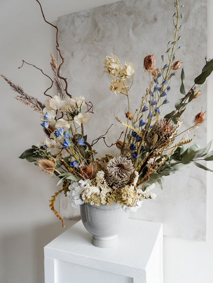 Dried flower arrangement in ceramic vase with natural toned florals accented with blue delphinium – close right side view.