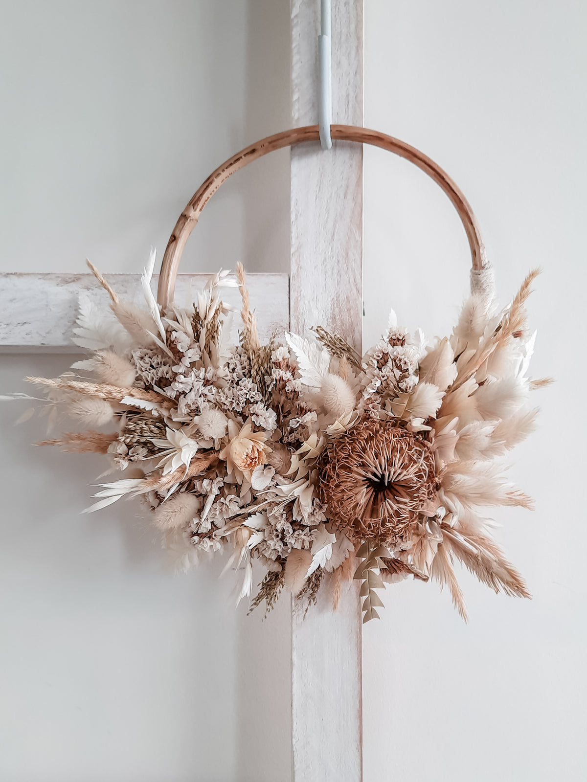 A rustic dried flower and grass wreath hanging on a white wooden ladder. The wreath features a natural rattan hoop, beige and cream dried grasses, white dried flowers, and a textured brown banksia.