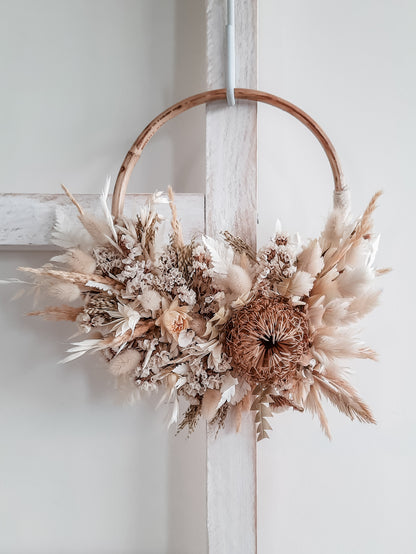 A rustic dried flower and grass wreath hanging on a white wooden ladder. The wreath features a natural rattan hoop, beige and cream dried grasses, white dried flowers, and a textured brown banksia.