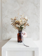 Load image into Gallery viewer, Dried flower arrangement in amber bottle vase with neutral white florals - distant full view.
