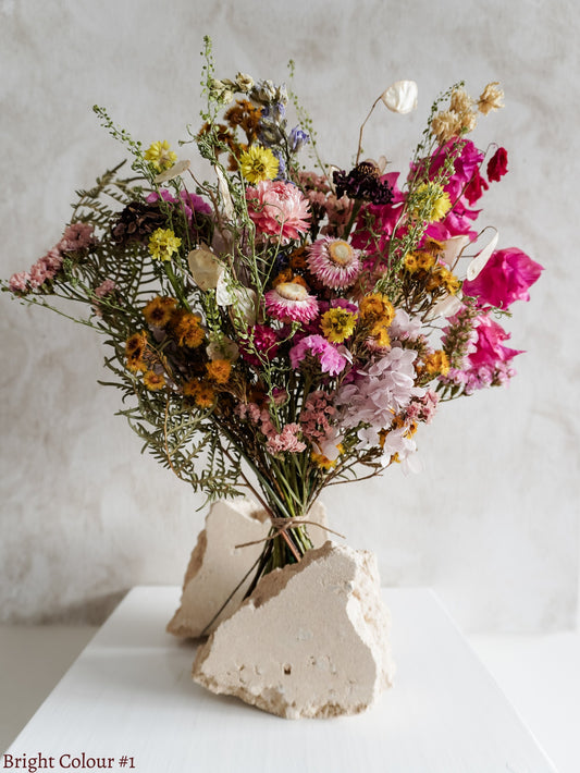 A dried flower bouquet featuring bright pink, mauve, and yellow flowers against a textured light gray background.