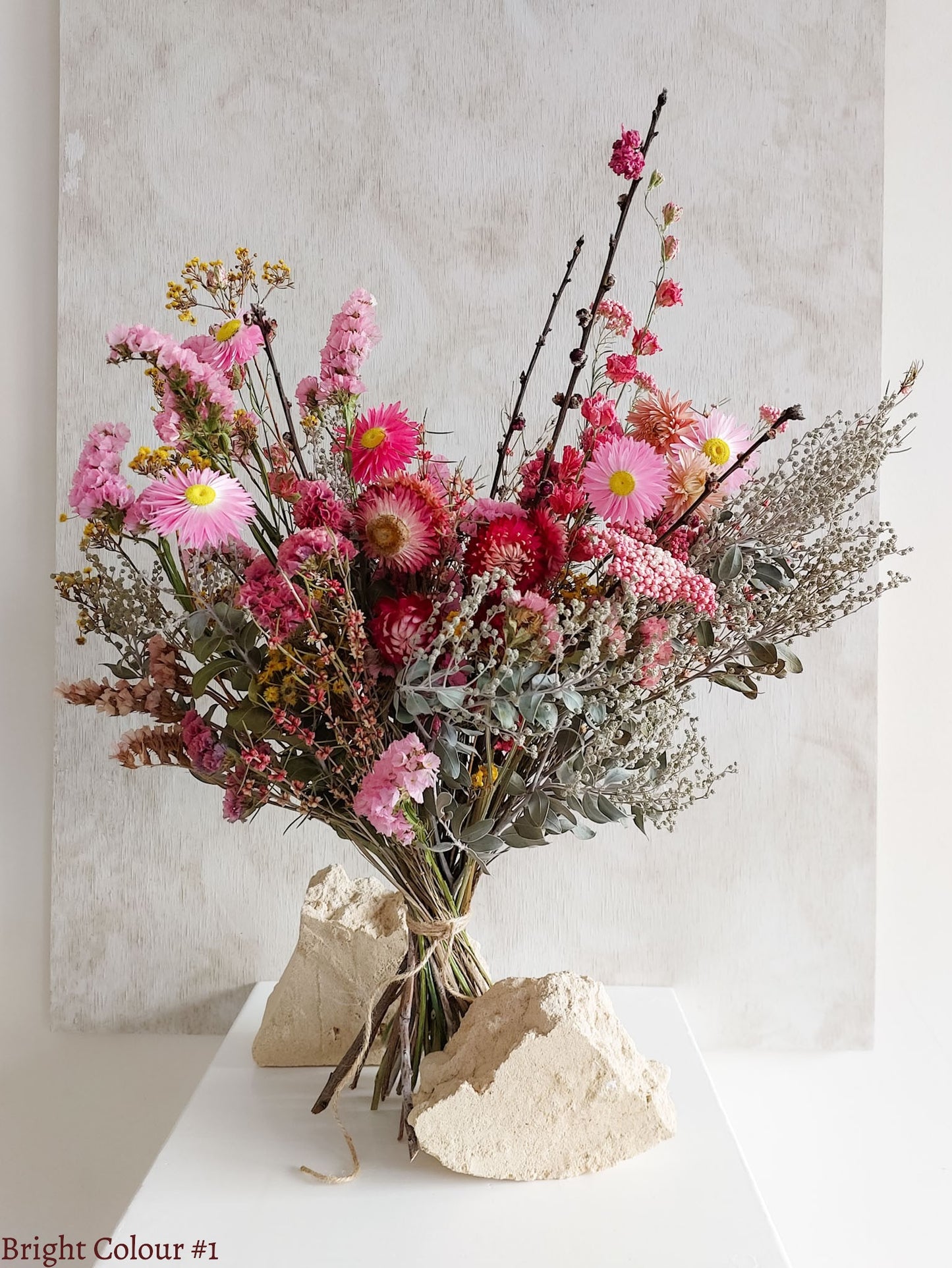 A dried flower bouquet featuring vibrant pinks and reds with sage green foliage, displayed on a white plinth against a textured gray background.