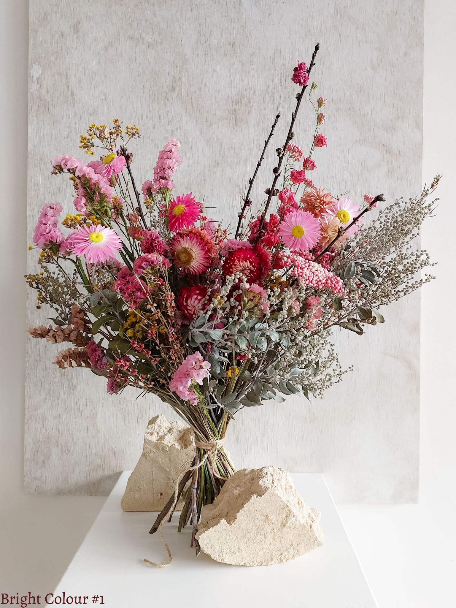 A dried flower bouquet featuring vibrant pinks and reds with sage green foliage, displayed on a white plinth against a textured gray background.