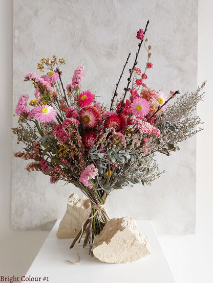A dried flower bouquet featuring vibrant pinks and reds with sage green foliage, displayed on a white plinth against a textured gray background.