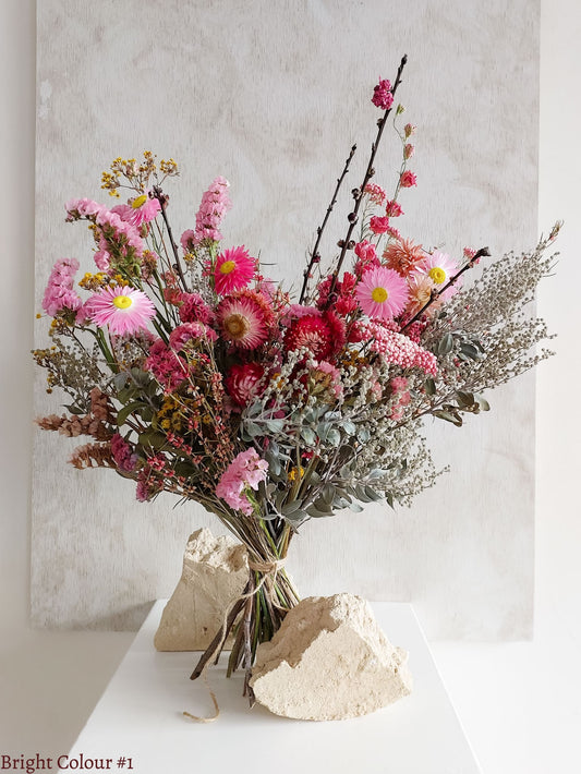 A dried flower bouquet featuring vibrant pinks and reds with sage green foliage, displayed on a white plinth against a textured gray background.