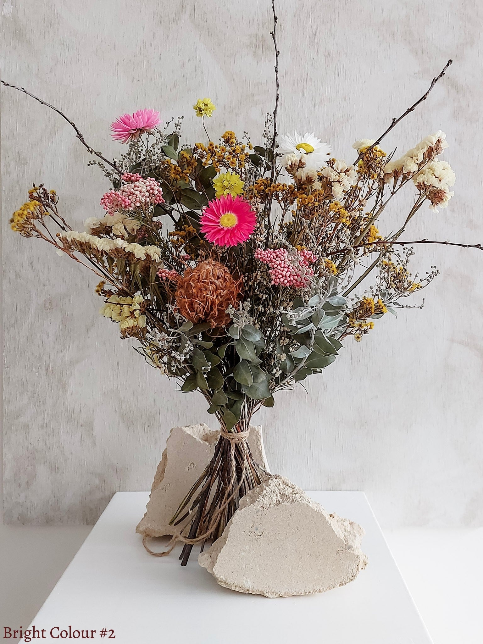 A dried flower bouquet featuring pink, yellow, and white flowers against a textured light gray background.
