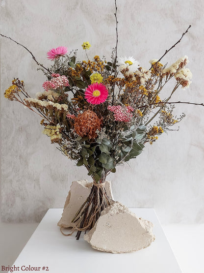 A dried flower bouquet featuring pink, yellow, and white flowers against a textured light gray background.