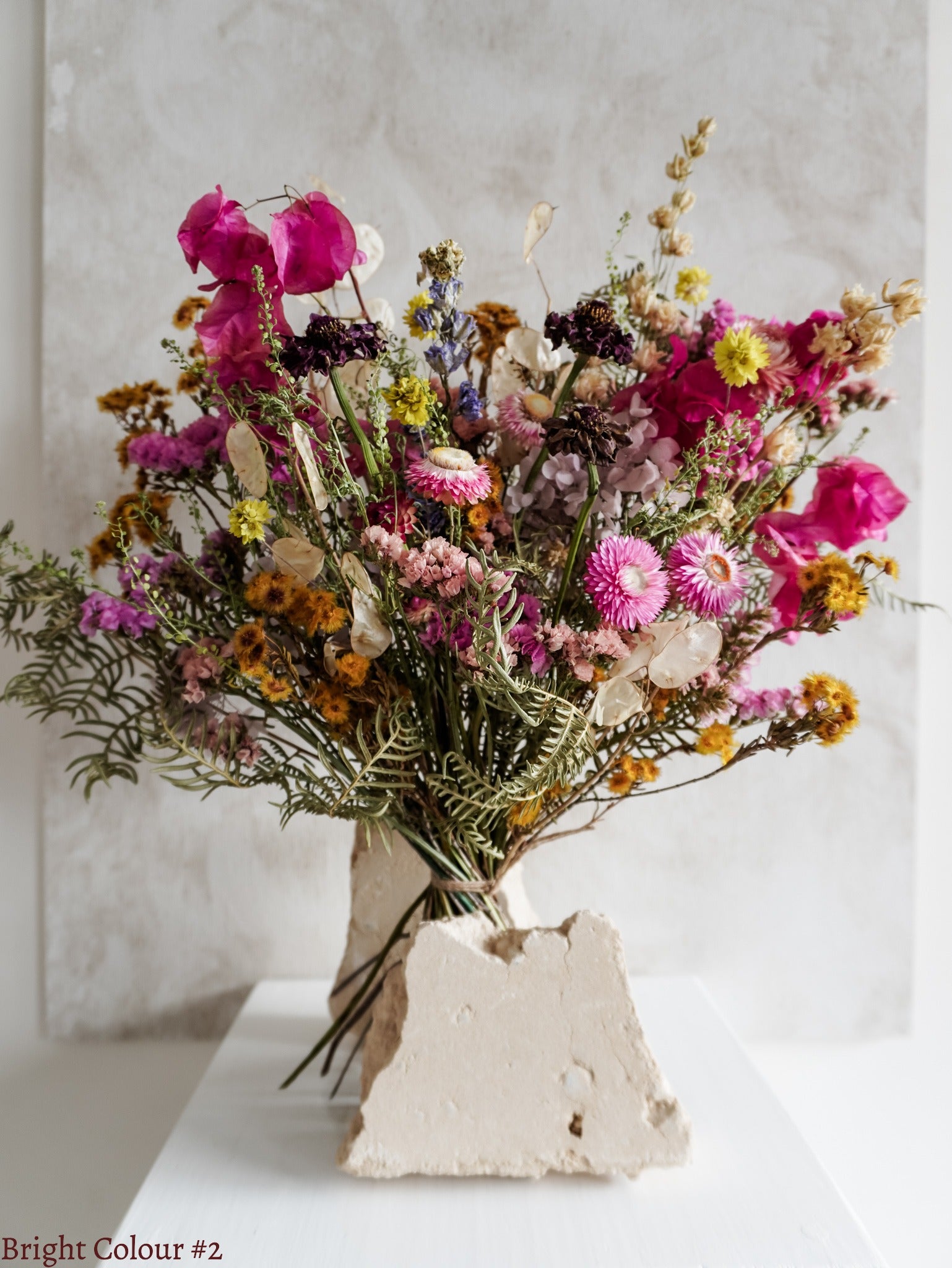 A dried flower bouquet featuring vibrant pink, yellow, and purple flowers with spring green foliage, displayed on a white plinth against a textured gray background.
