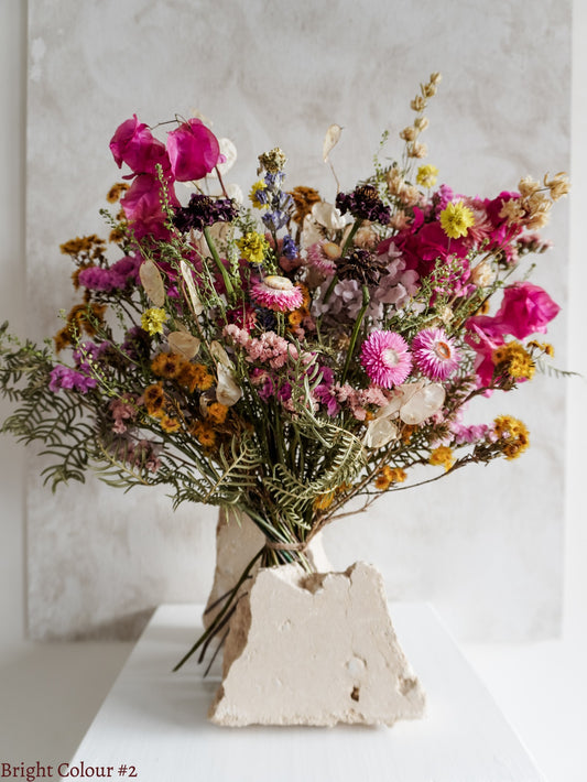 A dried flower bouquet featuring vibrant pink, yellow, and purple flowers with spring green foliage, displayed on a white plinth against a textured gray background.