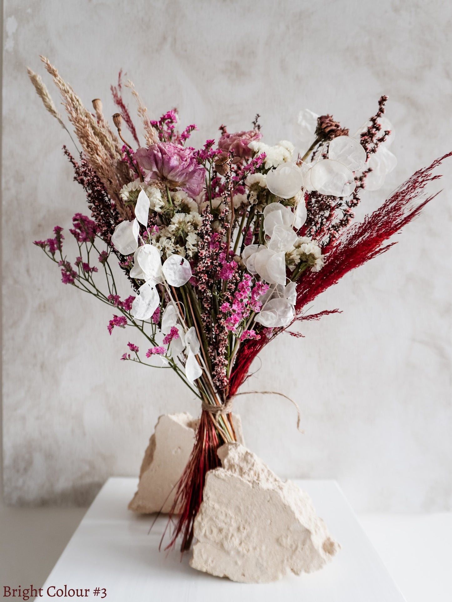 A dried flower bouquet featuring pink, white, and burgundy flowers against a textured light gray background.
