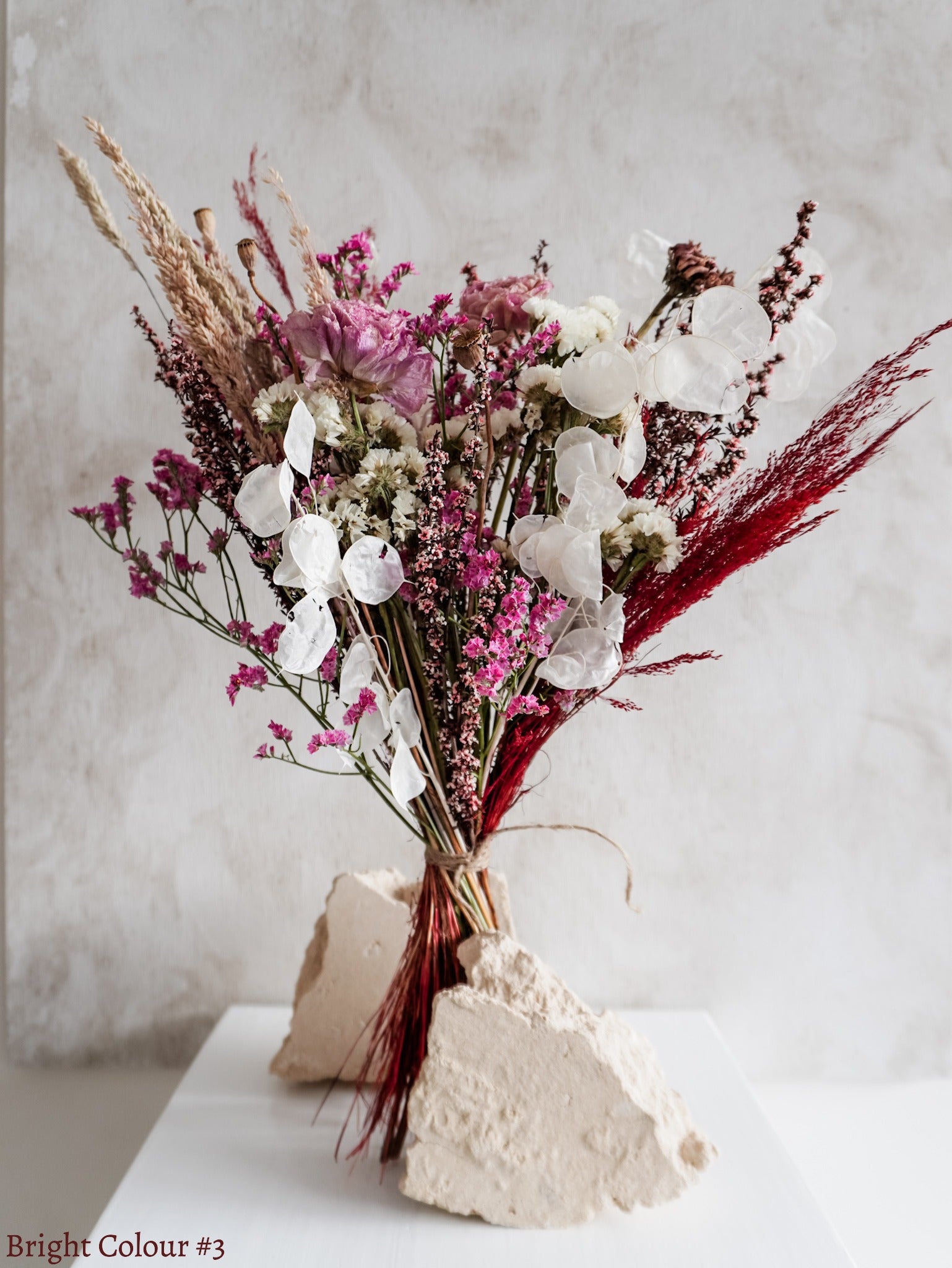A dried flower bouquet featuring pink, white, and burgundy flowers against a textured light gray background.