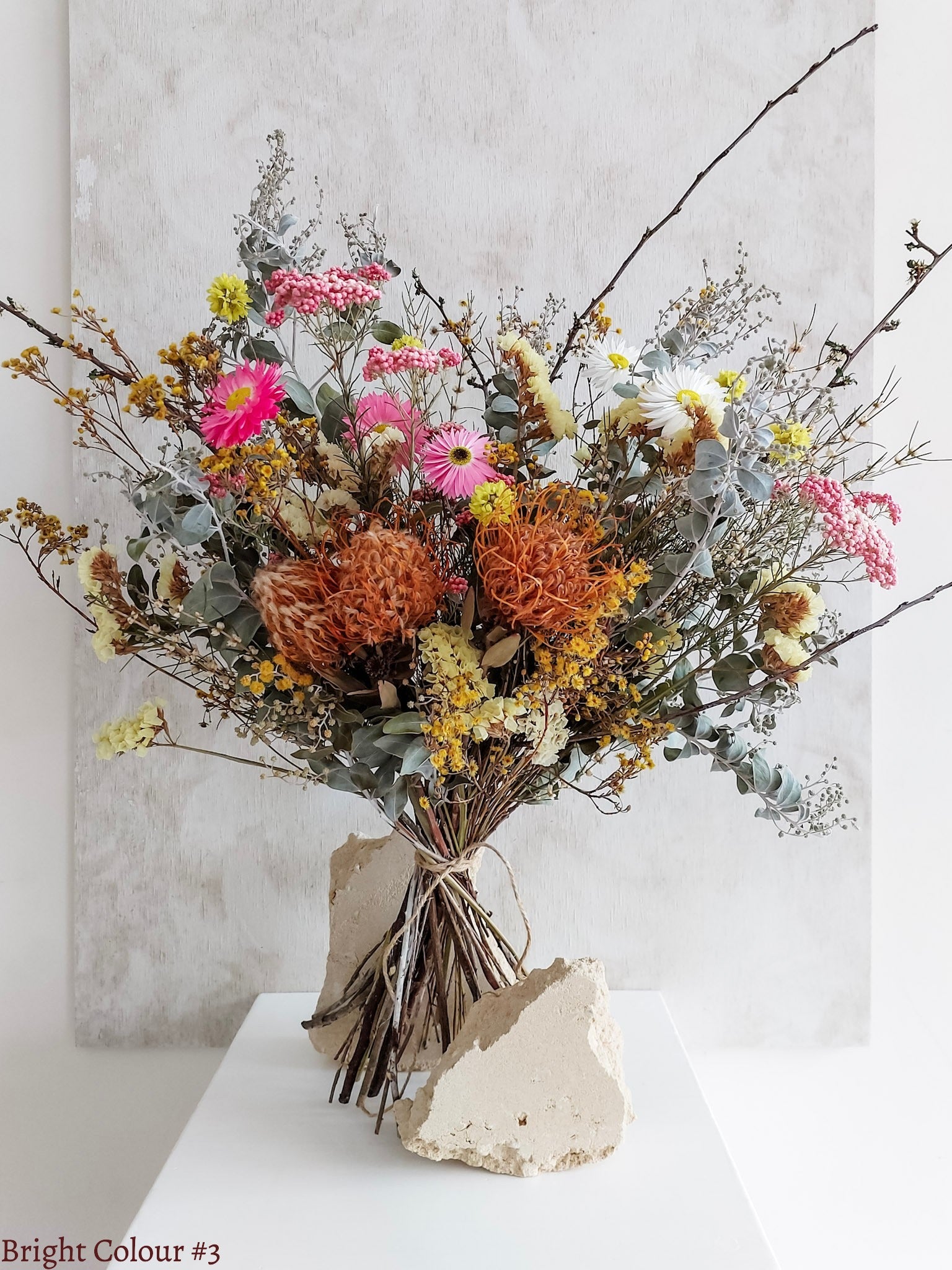 A dried flower bouquet featuring pink, yellow, and orange flowers with soft green foliage, displayed on a white plinth against a textured gray background.