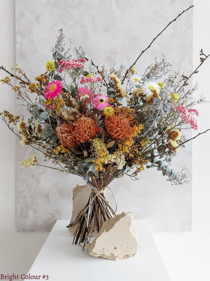 A dried flower bouquet featuring pink, yellow, and orange flowers with soft green foliage, displayed on a white plinth against a textured gray background.