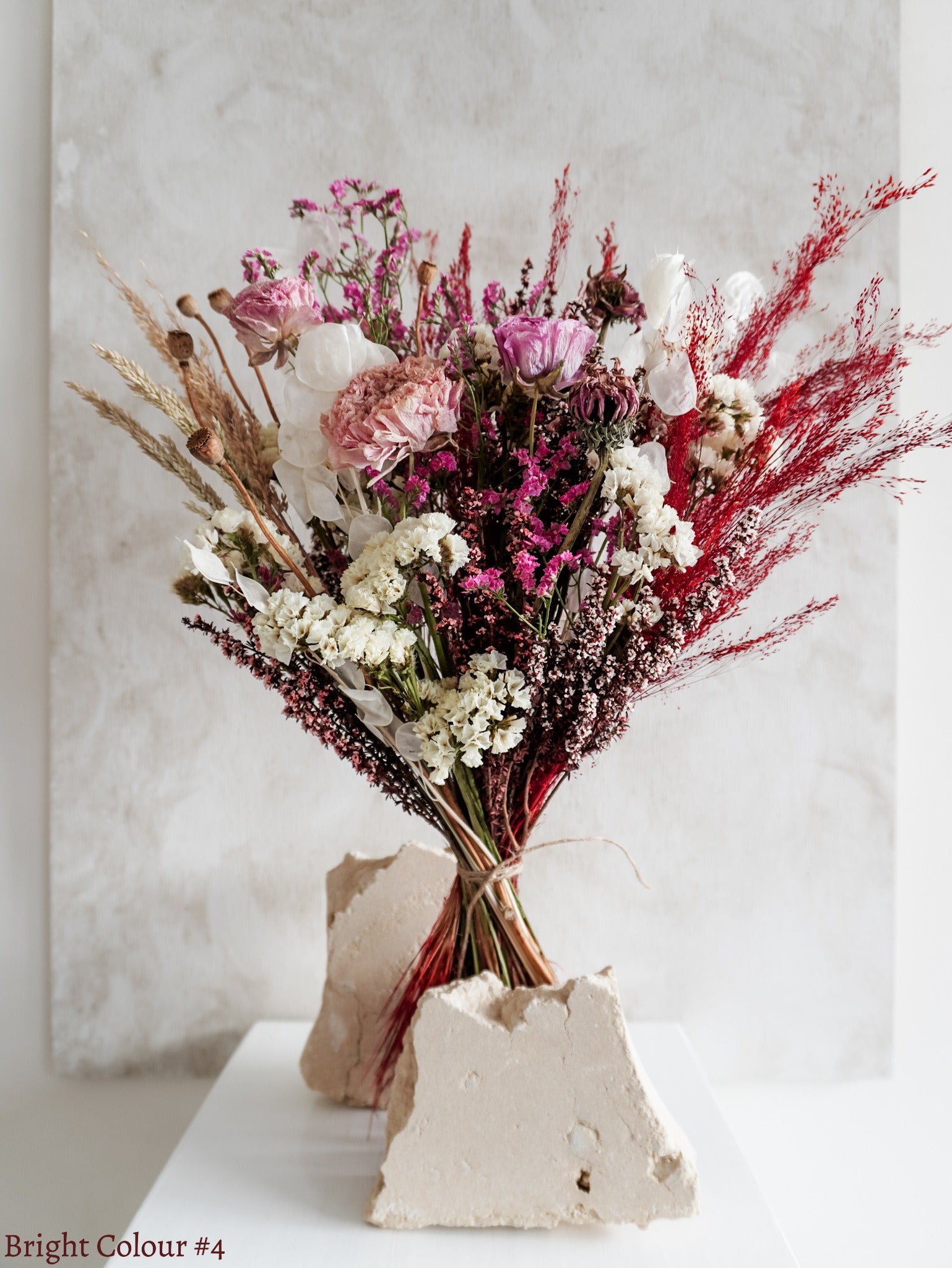A dried flower bouquet featuring vibrant pink, red, white and purple flowers, displayed on a white plinth against a textured gray background.