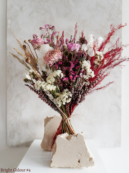 A dried flower bouquet featuring vibrant pink, red, white and purple flowers, displayed on a white plinth against a textured gray background.