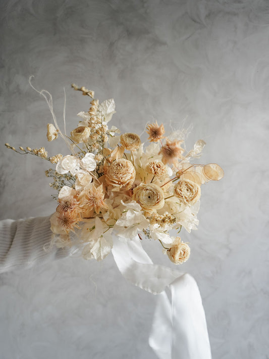 A dried flower bridal bouquet in soft beige and cream tones with a white ribbon, held by a hand wearing a white ribbed sweater against a textured light gray background.
