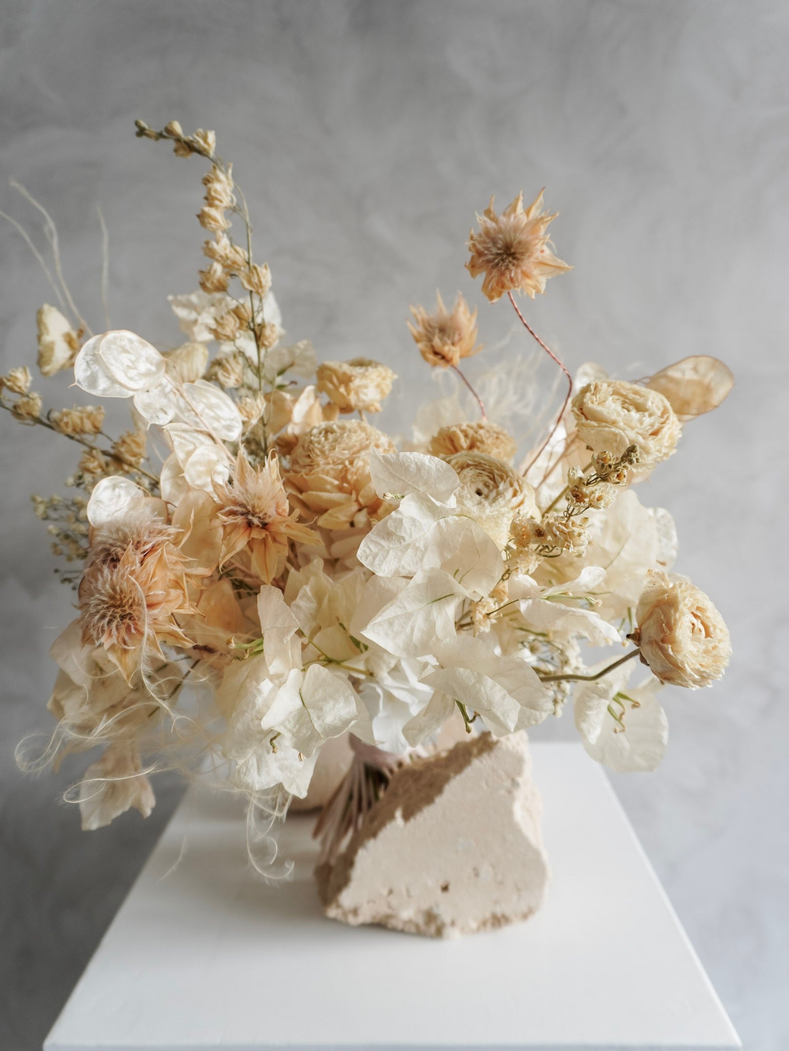 A dried flower bridal bouquet positioned against a rough, natural stone, featuring cream and soft beige florals, displayed on a white plinth against a textured gray background.