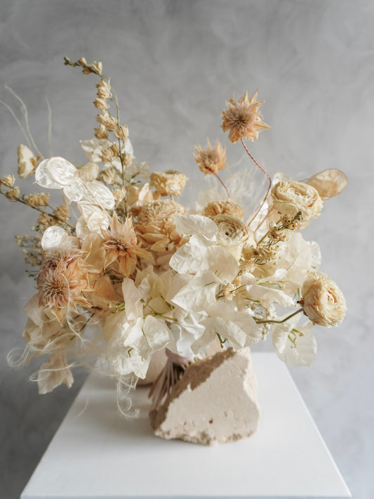 A dried flower bridal bouquet positioned against a rough, natural stone, featuring cream and soft beige florals, displayed on a white plinth against a textured gray background.